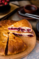 The round pie is cut into pieces. Inside, the filling is made of cherries and sugar. On a wooden table there are dishes, a cup of tea. Berries and flowers are laid out.