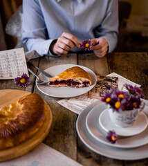 The hands of a girl in a blue shirt hold a cup of tea and a piece of cherry pie. There are plates and cups on a rustic table nearby. Berries and flowers are laid out. Sheets of musical notes.
