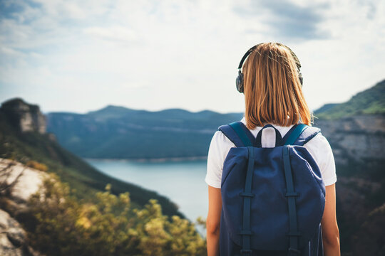 back view girl backpacker dreams listening to music in headphones relaxation on top rocky mountains, blonde hipster with backpack leisure after walking lake valley or green summer hills, empty space