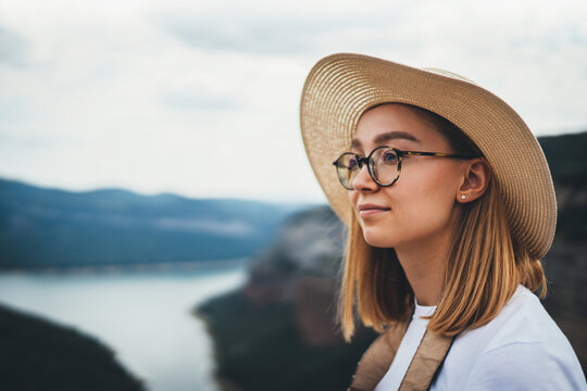 portrait of brooding young girl with blonde hair in straw summer hat and hipster glasses on backdrop river natura mountain landscape and blue sky, empty space for your design