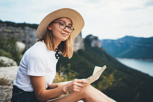 young blonde tourist holds in hands map of  landscape and looks views top of mountain, cute smiling girl in hat and glasses leisure in nature outdoors, concept of travel vacation