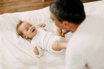 Fototapeta na wymiar Happy father playing with adorable baby in the bedroom in a snow-white bed