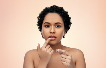 beauty, make up and cosmetics concept - beautiful young african american woman applying lip gloss with finger over beige background