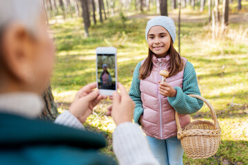 picking season, leisure and people concept - happy smiling grandmother with smartphone photographing granddaughter with mushrooms in basket in forest
