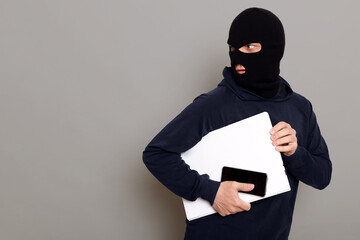 Man steals laptop and phone, runs away and turns back, being of persecution, wears robber mask and black turtleneck, advertising space, isolated over gray background. - obrazy, fototapety, plakaty