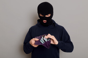 Surprised man thief dressed in robber mask holding open wallet in his hands, shocked by what he sees, steals big sum of money, posing isolated over gray background.