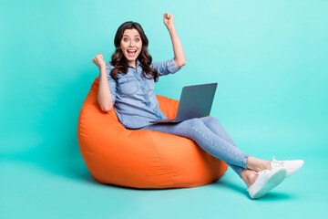 Photo of lucky funny young woman wear jeans shirt sitting beanbag chatting modern gadget rising fists isolated turquoise color background