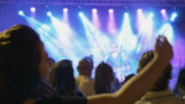 Young people clap their hands in front of the stage. Blurred silhouettes of people are dancing to the rhythm of the music at an outdoor summer night concert.