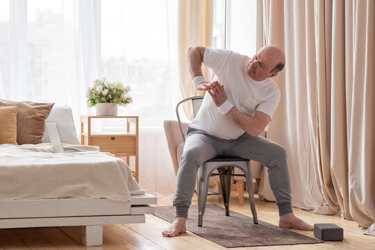 Senior Caucasian Man Stretching Side Sitting On Chair At His Living Room.