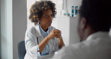 African-american woman doctor sitting at table and talking to male colleague in office - Powered by Adobe