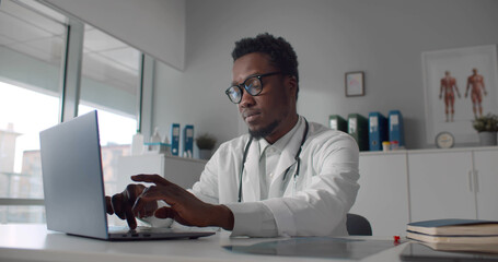 African american doctor wearing white coat typing on laptop in hospital office