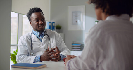 Afro-american male doctor talking to patient sitting at desk in office.