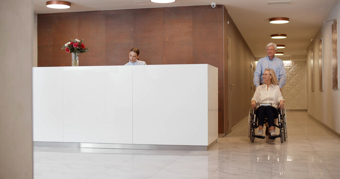 Mature man pushing wheelchair with wife and talking to staff at reception desk