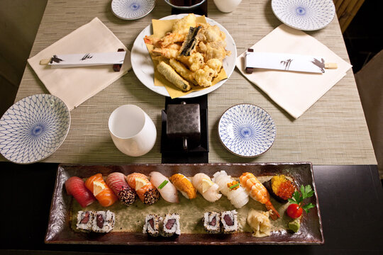 Sushi and Tempura in Japanese Food Restaurant – Gourmet Dishes, Ceramic Plates with Deep Fried Vegetables, Rows of Rolls and Nigiri, Red Caviar, Chopsticks, Black Wood Table and Placemats – Top View