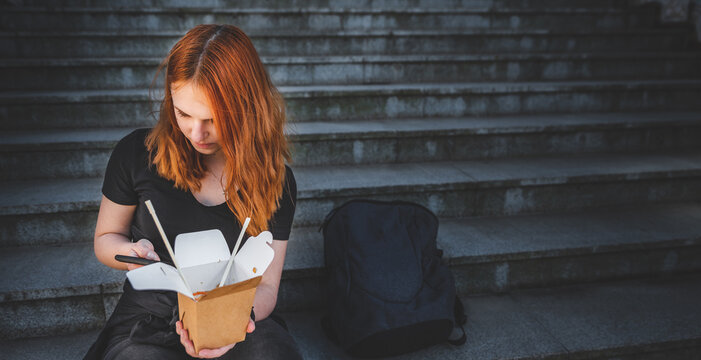 young woman or teenage girl eating asian fast food from takeaway box sitting on the steps on city street. Thai noodles in paper box takeaway street food