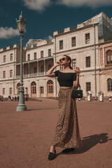 Elegant woman wearing black top, leopard skirt and sunglasses, holding black leather handbag, standing and posing at square against beautiful palace.