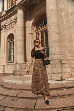 Elegant woman wearing black top, leopard skirt and sunglasses, holding black leather handbag, standing and posing at square against beautiful palace.