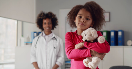 Portrait of little african girl holding teddy bear with young female doctor standing on background