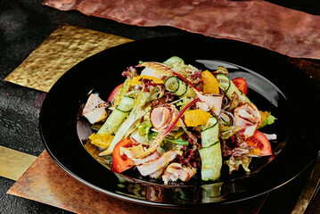 salad with ham, orange slices, tomato, cucumber and lettuce leaves in a black plate on a copper background. 