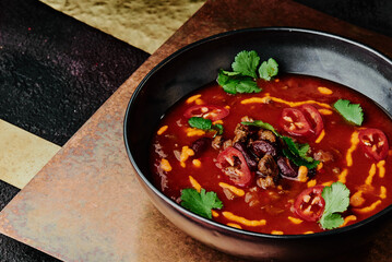 spicy tomato soup with beans, chili pepper, cheese and parsley in a black plate on a copper background