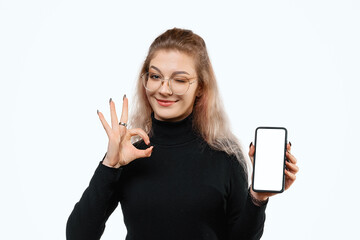 Positive young woman showing OK gesture with hand and holding empty smartphone screen, recommend...