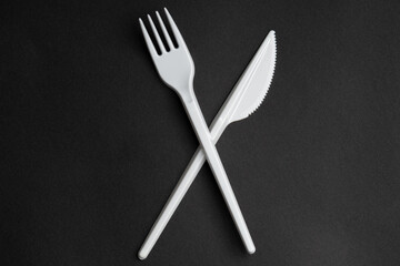 White fork and knife crosswise on black background. Plastic cutlery, ecology, environmental pollution by plastic, disposable tableware, waste recycling concept. copyspace