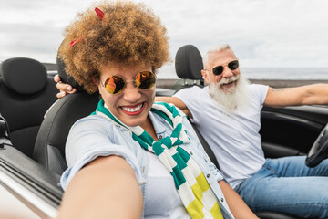 Trendy senior couple having fun taking a selfie with mobile phone in convertible car during summer vacation - Focus on mature woman face