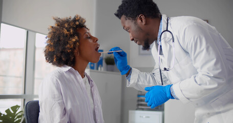 Afro-american male doctor examining young woman throat in office.