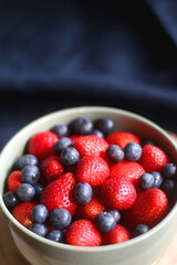 Bowl of blueberries and strawberries on wooden tray. Selective focus, dark textile background.