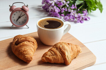 Wake up and good morning concept. Coffee with croissants, alarm clock and flowers on the table. Delicious breakfast, start of the day. Close-up, selective focus