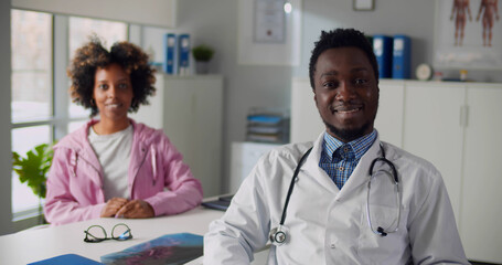 Fototapeta premium Portrait of afro-american male doctor and female patient smiling at camera in clinic office