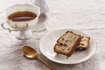 Slice of Pound Loaf Cake with raisins on white porcelain plate and cup of tea on greige linen tablecloth. Selective focus