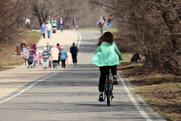 Girl riding on a bicycle on a path in a park on walking people background. Spring leisure, weekend activities, cycling concept