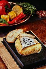 Grilled Camembert Cheese on a black square stone plate with toast and grilled vegetables on a copper background.