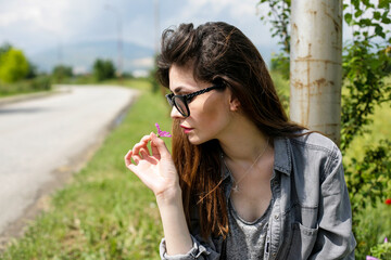 Young woman is sitting alongside the road and is smelling a flower.