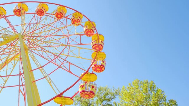 A colorful yellow and orange ferris wheel slowly rotates in an amusement park against a bright blue sky on a sunny summer day. The concept of entertainment and fun during the holiday, carnival