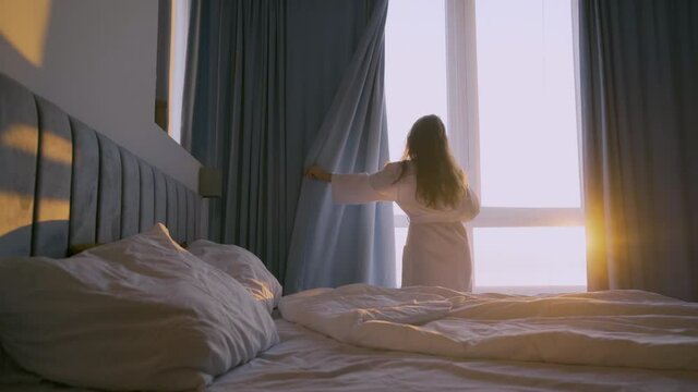 Young curly haired woman in white bathrobe opens curtains on large panoramic window looking outside in elegant hotel room at sunrise backside view