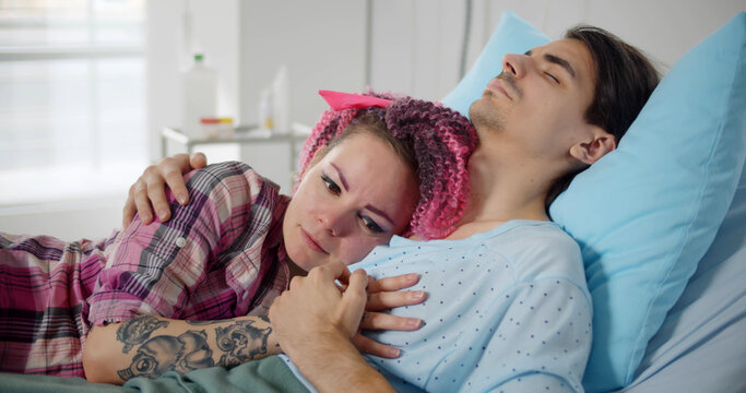 Worried and sad young woman hugging sick husband sleeping in hospital bed