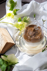 Delicious dalonga coffee with ice and milk in glass cup. Iced frothy Dalgona Coffee, a trendy fluffy creamy whipped coffee. Korean drink latte espresso with coffee foam. Green flowers and a book.