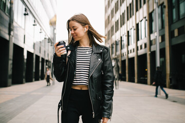 Young pretty girl listening to music in the big city. girl in a leather jacket with headphones drinking coffee near business center . smiling and loathing