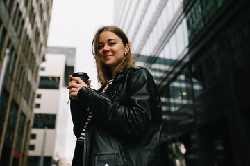 Young pretty girl listening to music in the big city. girl in a leather jacket with headphones drinking coffee near business center . smiling and loathing