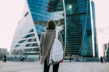 A blonde young girl with a white backpack dressed in a jacket stands against the background of...
