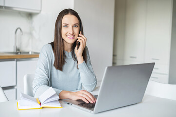 Smiling and positive young woman talks on smartphone working with laptop from home, female employee holding phone connection with customers or colleagues by mobile phone, looks at the camera