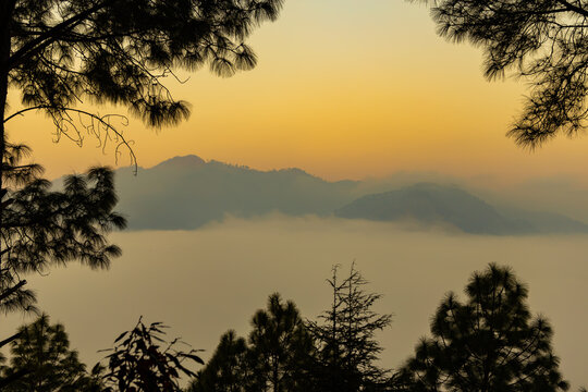 A view of mountains and valley covered in early morning mist and the orange glow from sunlight with trees framing the corners of the image