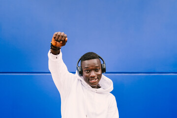 Young African American black male listening music with wireless headphones while doing protest expression showing fist in the air. Wearing a white sweatshirt over a blue wall. BLM