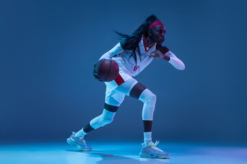 Obraz premium Beautiful african-american female basketball player in motion and action in neon light on blue background. Concept of healthy lifestyle, professional sport, hobby.