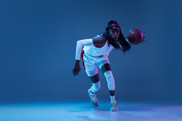 Beautiful african-american female basketball player in motion and action in neon light on blue background. Concept of healthy lifestyle, professional sport, hobby.