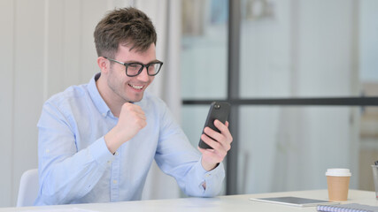 Young Man Celebrating Success on Smartphone in Office 