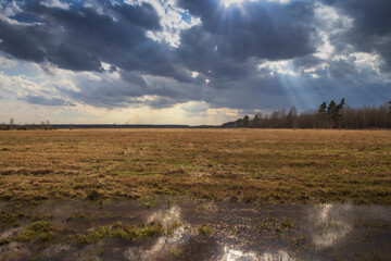 Early spring in the field. Sunlight through the clouds. Forest on the horizon. Dramatic clouds. Landscape with a field and trees. The sun shines with bright rays.
