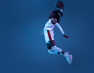 Beautiful african-american female basketball player in motion and action in neon light on blue background. Concept of healthy lifestyle, professional sport, hobby.